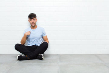 young handsome man feeling confused, puzzled and insecure, pointing to self wondering and asking who, me? sitting on cement floor