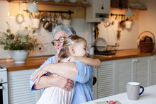 Grandmother Hugging Child In Cozy Kitchen At Home. Happy Family Giving Thanks. Cute Little Girl And Senior Woman Is Enjoying Kindness Together. Lifestyle Moment. Holiday Thanksgiving.