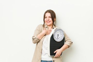 Wall Mural - Young curvy woman holding a scale laughs out loudly keeping hand on chest.