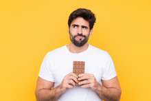 Young Handsome Man With Beard Over Isolated Yellow Background Taking A Chocolate Tablet And Having Doubts