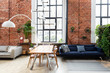 Industrial interior of living room in spacious loft apartment with big windows. Indigo sofa with pillows, modern table with chairs and stylish lamp in comfortable lounge. Brick wall. 