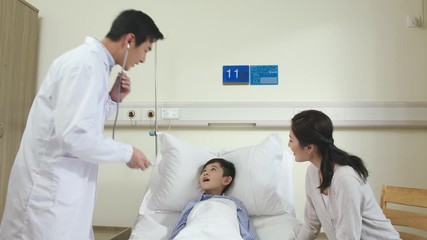 Wall Mural - young asian male doctor making rounds examining and talking to child patient and mother in hospital ward