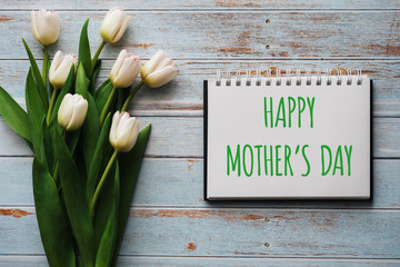 Wall Mural - Bouquet of white flowers of tulips on the background of blue boards with a notebook with lettering Happy Mother's Day