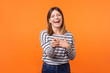 © khosrork - Portrait of amusing joyous young woman with brown hair in long sleeve striped shirt closing eyes, pointing finger and laughing out loud at funny joke. indoor studio shot isolated on orange background