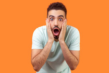 Unbelievable Shocking News! Portrait Of Stunned Brunette Man With Beard In Casual White T-shirt Holding Face And Keeping Mouth Wide Open In Amazement. Indoor Studio Shot Isolated On Orange Background