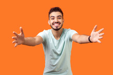 Come into my arms. Portrait of good natured, extremely happy brunette man with beard in white t-shirt reaching out to camera, stretching arms to hug you. studio shot isolated on orange background
