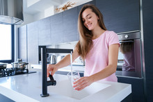 Happy Attractive Joyful Brunette Woman Pours Fresh Clean Filtered Purified Water For Drinking From A Tap Into A Glass At Kitchen At Home. Healthy Lifestyle And Quench Thirst