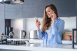 Young happy woman using smartphone and white wireless earphones for listening music and audio book in the kitchen at home. Modern mobile people