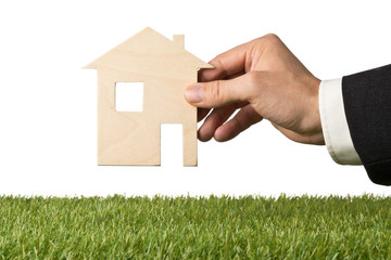 Wall Mural - Business man holding little wooden miniature house model over green grass isolated on white background with copy space - ecological living or house building concept