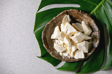 Wall Mural - Chopped pieces of raw organic cocoa butter in craft ceramic plate on green exotic monstera leaf over grey spotted background. Flat lay, copy space