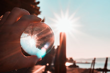 Hand Holding A Crystal Glass Ball, Morning Harsh Sun In The Sky Reflected In The Ball. Outlines Of Beach In The Background