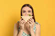 Young woman eating tasty sandwich on yellow background