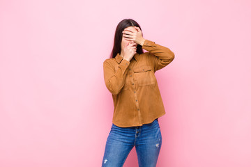 young pretty woman covering face with both hands saying no to the camera! refusing pictures or forbidding photos against pink wall