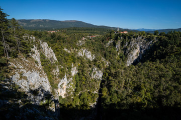 View of Unesco world heritage site Skocjan caves (Skocjanske jame). Looking to a deep gorge with cave entrance, sinkholes and walking paths. Village of Skocjan on a rock in the background.