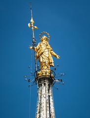 Wall Mural - Madonnina atop Milan Cathedral at the height of 108.5 m in Milan, Italy. Golden statue of Madonna close-up on the blue sky background.