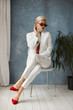 Beautiful young woman with perfect blond hair in elegant white suit and red shoes posing in studio. Fashionable blonde model girl in stylish outfit and sunglasses. Lady boss ready for office meeting