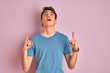 Teenager boy wearing casual t-shirt standing over blue isolated background amazed and surprised looking up and pointing with fingers and raised arms.