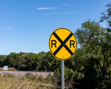 Yellow Railroad Crossing Warning Sign On Rural Road With Trees And Train Tracks In Background