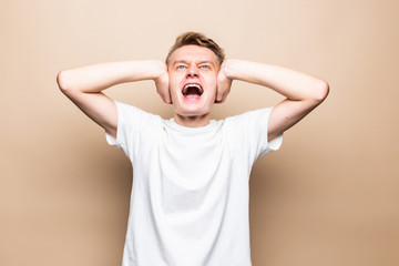 Young man covering his ears with hands opens his mouth screaming isolated on a beige background.