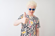 © Krakenimages.com - Young albino blond man wearing colorful t-shirt and sunglasses over isolated red background doing happy thumbs up gesture with hand. Approving expression looking at the camera with showing success.