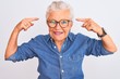 Senior grey-haired woman wearing denim shirt and glasses over isolated white background smiling pointing to head with both hands finger, great idea or thought, good memory