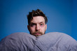 © phoenix021 - Serious man on blue background holding head on pillow and looking at camera, having trouble sleeping concept