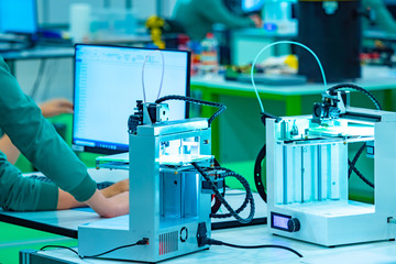 Poster - Two 3d printers and a computer are on the table. The young man sets up printing on a 3d printer. Devices for printing three-dimensional images. The machine prints the specified objects in layers.