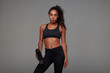 © timtimphoto - Studio shot of attractive young slim dark skinned lady with curly long brown hair relaxing after workout in gym, posing over grey background with fitness bottle in her hand