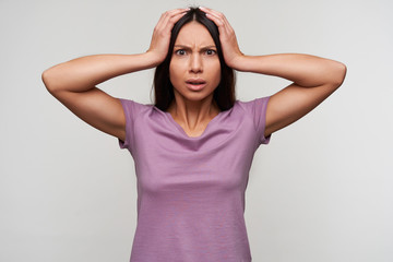 Bewildered young pretty brown-eyed brunette lady clutching her head and looking confusedly at camera, frowning her face while posing over white background in purple t-shirt