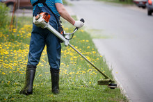 View From The Back To The Man With A Gasoline Mower. Worker In Overalls Mows The Grass On The Lawn With A Trimmer Near The Roadway.