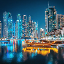 Multicolored Lights Of The Night City In The Dubai Marina District. Stylized Ancient Arabic Ship Abra Dhow With Lights In The Foreground.