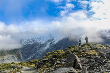 Hikers Taking In View Surrounded By Glacial Mountains In The Clouds.