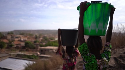 back view of two young african girls carrying water buckets down the hill