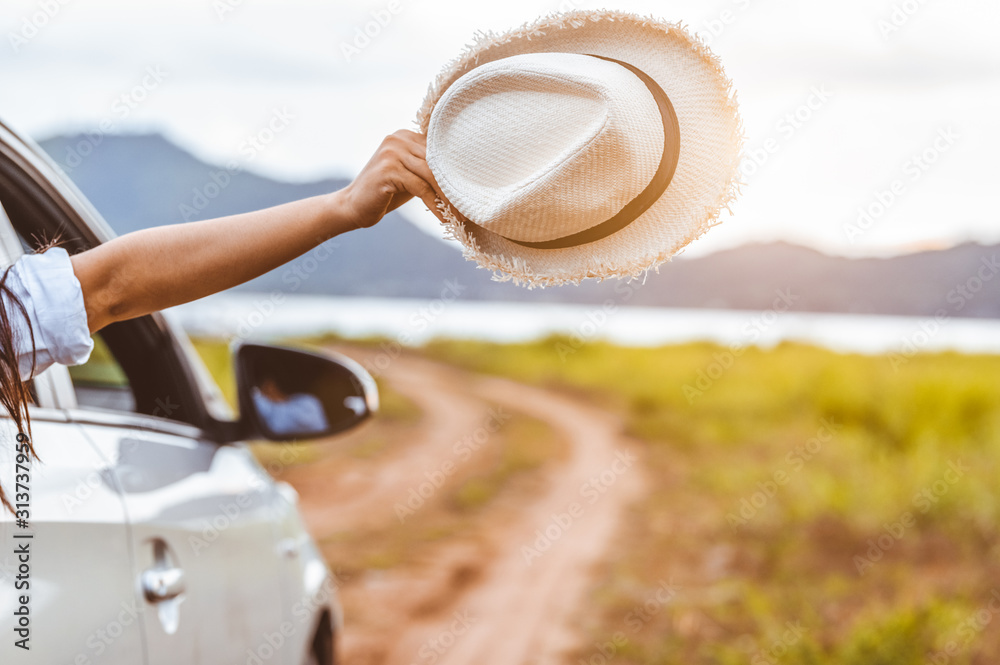 Happy woman hand holding hat outside open window car with meadow and mountain lake background. People lifestyle relaxing as traveler on road trip in holiday vacation. Transportation and travel concept - obrazy, fototapety, plakaty 
