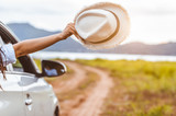 Fototapeta  - Happy woman hand holding hat outside open window car with meadow and mountain lake background. People lifestyle relaxing as traveler on road trip in holiday vacation. Transportation and travel concept