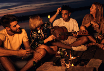 Group of young friends enjoying at the lake at night. They sitting around the fire singing and having fun at camping.