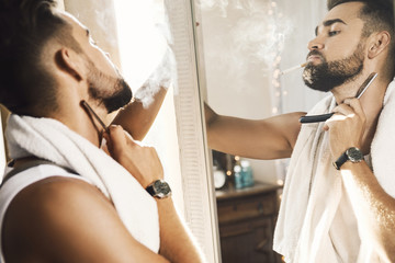 Wall Mural - Man looking in the mirror, smoking a cigarette and shaving his beard with a straight razor