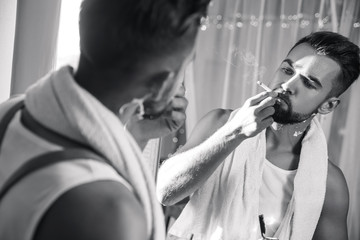 Wall Mural - Man looking in the mirror, smoking a cigarette and shaving his beard with a straight razor