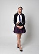 © faestock - full length portrait of a pretty brunette girl wearing a school uniform of black jacket and plaid skirt. Standing pose isolated and holding books, against a studio background.