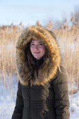 Medium vertical portrait of pretty smiling blonde young woman in winter coat with faux fur trim hood standing outside in front of ornamental grasses during a sunny winter afternoon