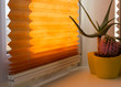 Pleated blinds with orange folded fabric on the window close up. On the windowsill stands home plant in yellow flower pot. Cordless pleated shade with white lower bar.