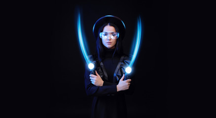 Beautiful woman in futuristic black dress over dark background. Gamer girl in glasses of virtual reality with controllers in hands. Augmented reality, game, hobby concept. VR. Blue neon light.