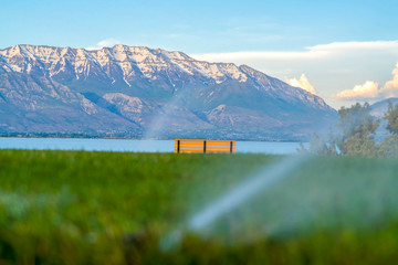 Wall Mural - Bench overlooking lake and mountain with blurred sprinkler watering green grass