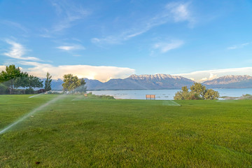 Wall Mural - Sprinklers on green grassy field with scenic lake and snowy mountain background