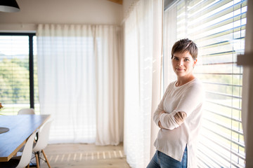 Portrait of young woman standing by window indoors at home, looking at camera.