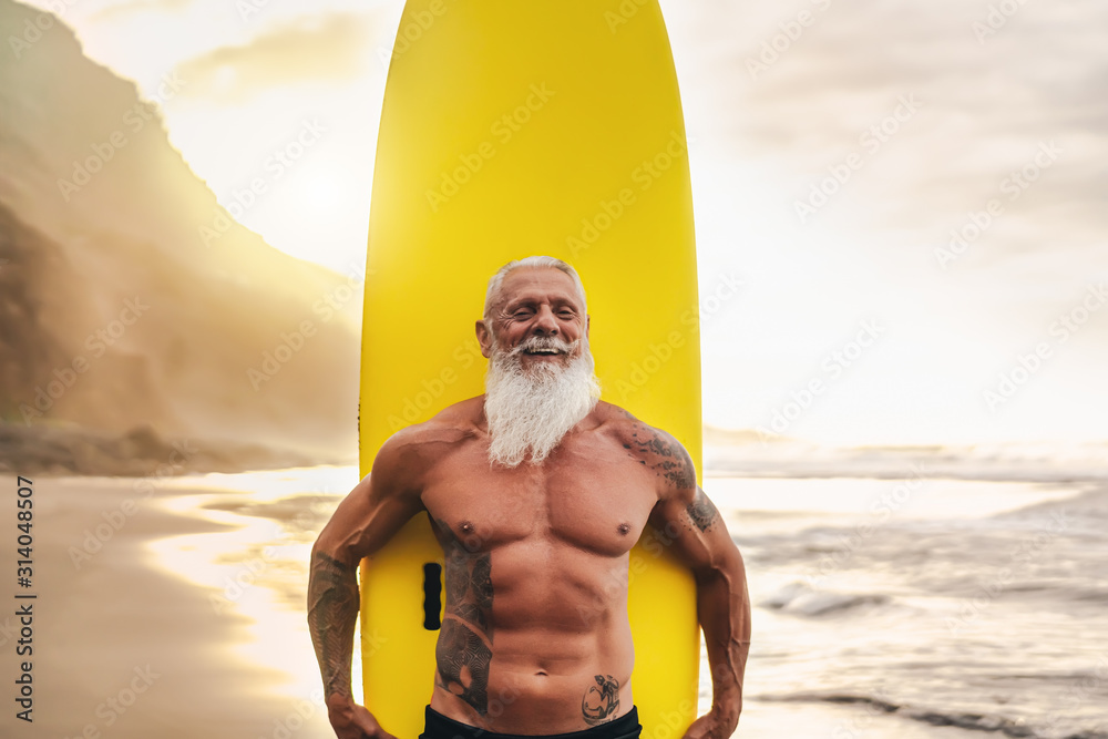 Happy fit senior having fun surfing at sunset time - Sporty bearded man training with surfboard on the beach - Elderly healthy people lifestyle and extreme sport concept - obrazy, fototapety, plakaty 