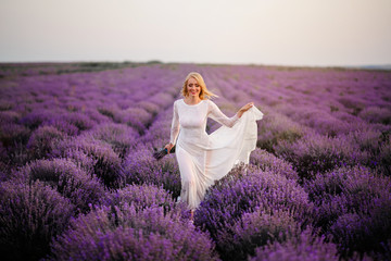 Happy smiling bride dressed in boho style dress walks on a blooming lavender field at sunset.