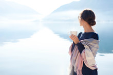 Woman Drinks Morning Coffee At Sea Beach. Cozy Winter Picnic. Girl Enjoying Calm Nature, Travel, Relaxation, Wellbeing. Blue Background Of Mountains, Still Water. Copy Space. Rear View Female Portrait