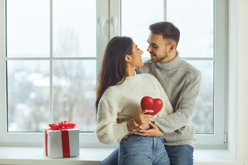 Valentine's day. Beautiful smiling couple gives a heart on a background of a window in a room