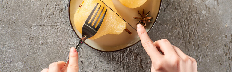 cropped view of woman eating delicious pear in wine with fork and knife on grey concrete surface, panoramic shot